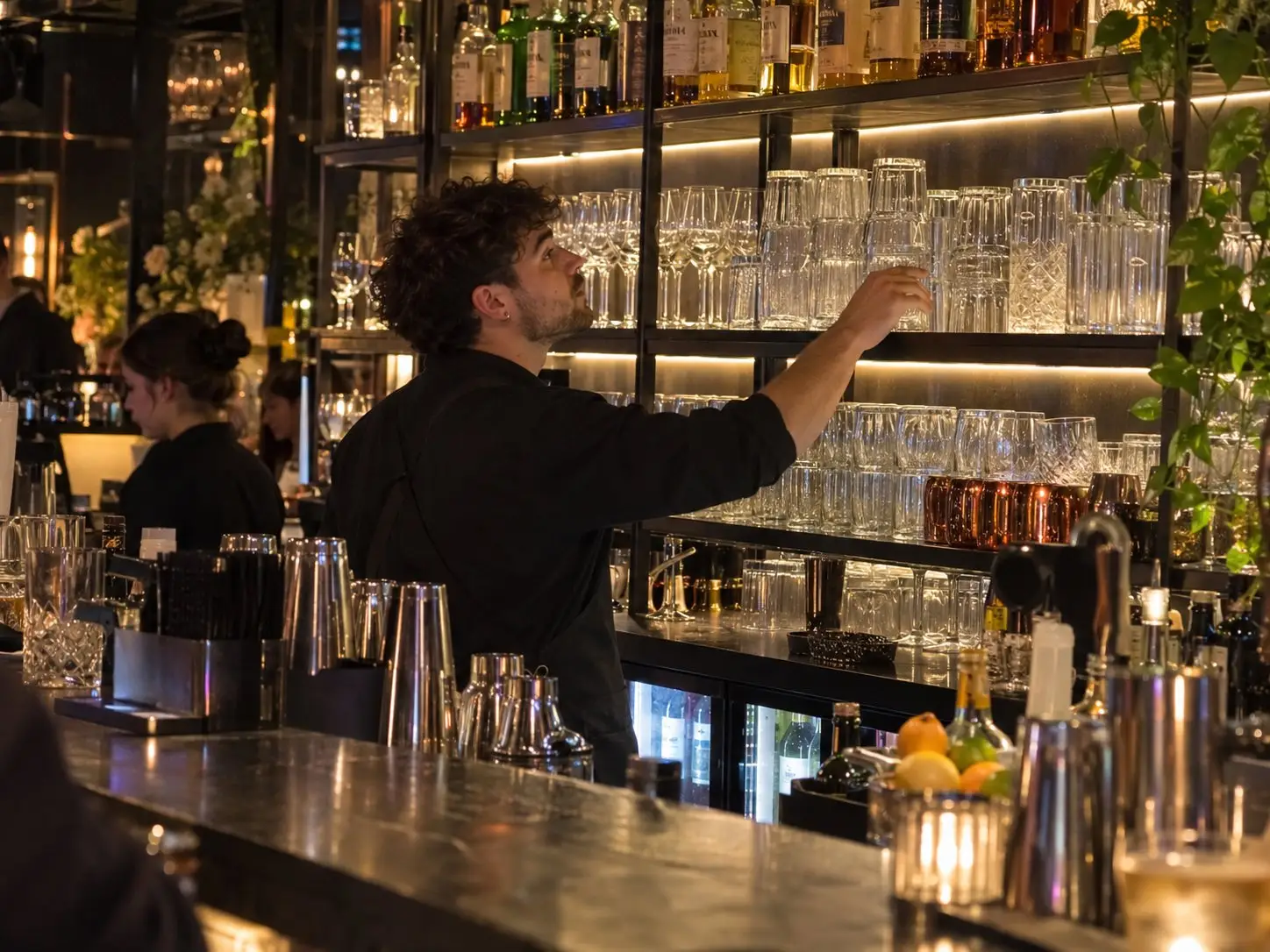 Bartender puts freshly washed glasses back on the shelf of a lit back bar
