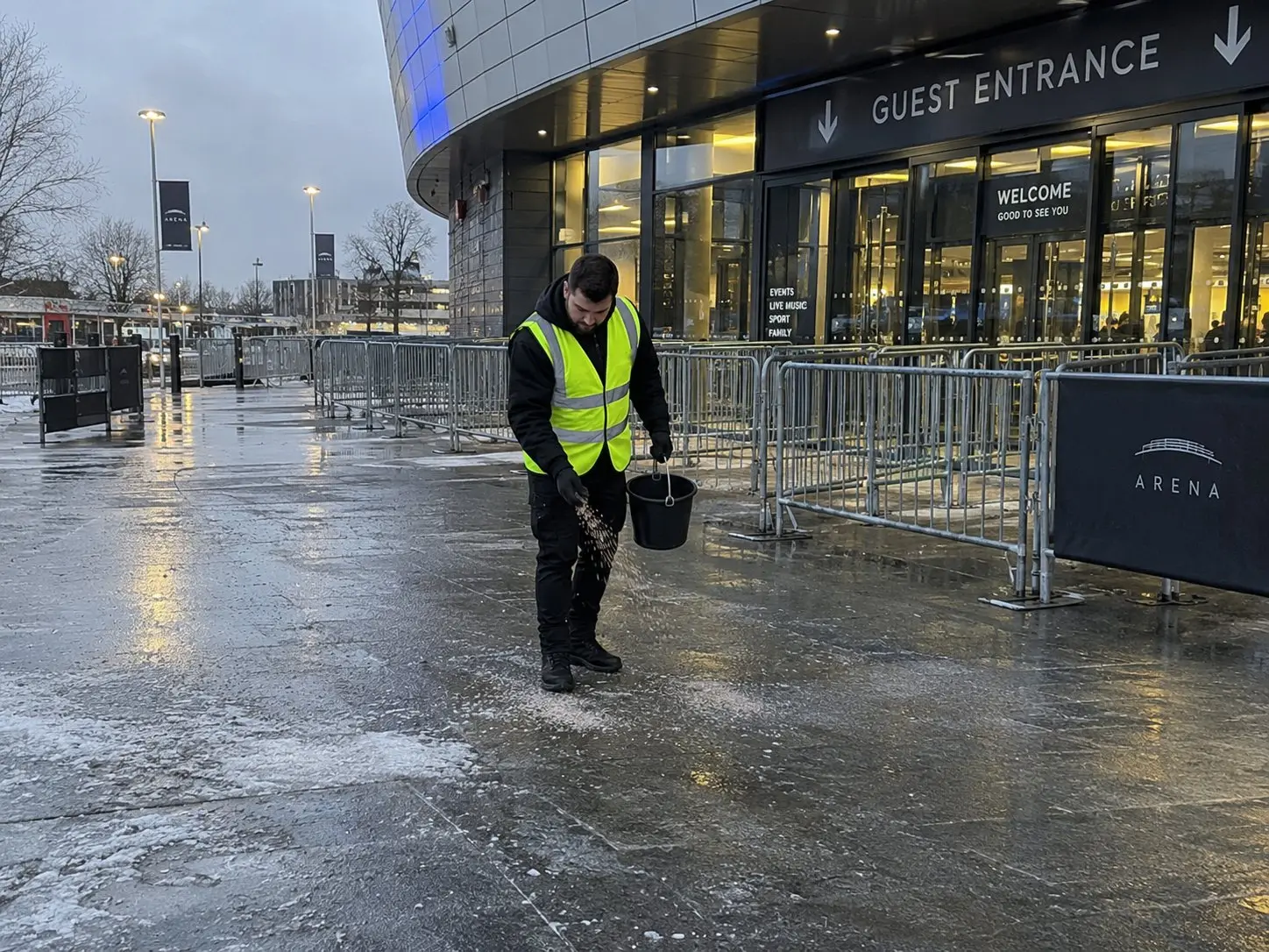 Eventhelfer in Warnweste streut vor dem Gästeeingang einer Arena Salz auf den vereisten Vorplatz