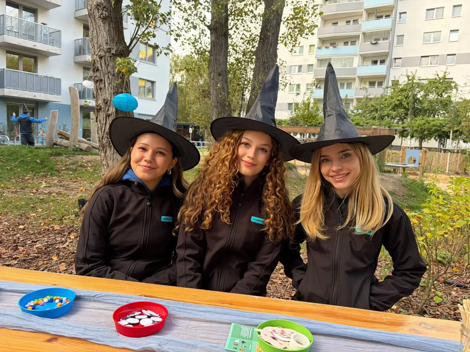 Three staff members in HOWOGE jackets and witch hats at the craft table of a courtyard festival