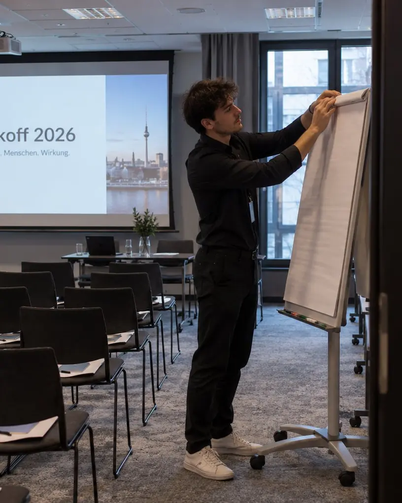 Servicekraft bereitet im Workshop-Raum einer Kick-off-Tagung das Flipchart vor, im Hintergrund Leinwand und Stuhlreihen – Eventpersonal in Berlin