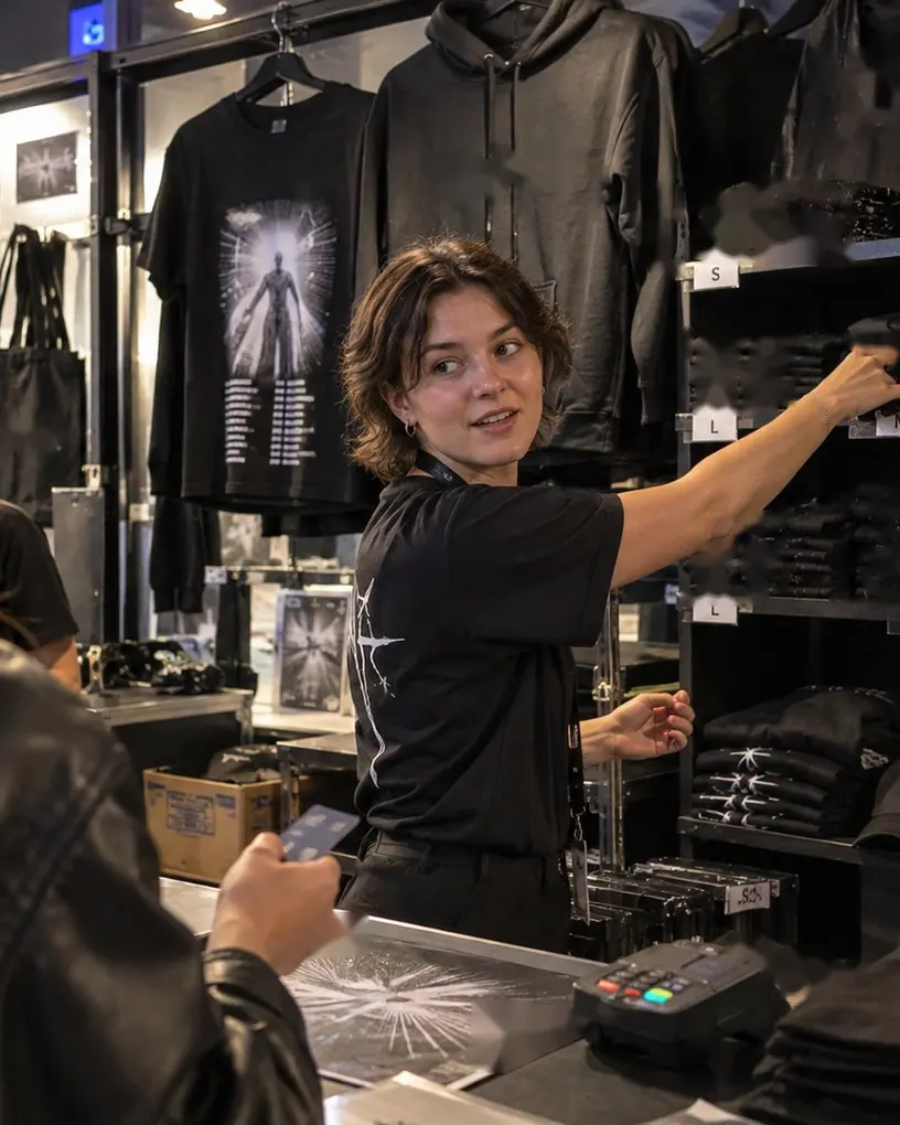 Verkäuferin am Merch-Stand greift ein T-Shirt aus dem Regal, eine Kundin hält ihre Karte bereit