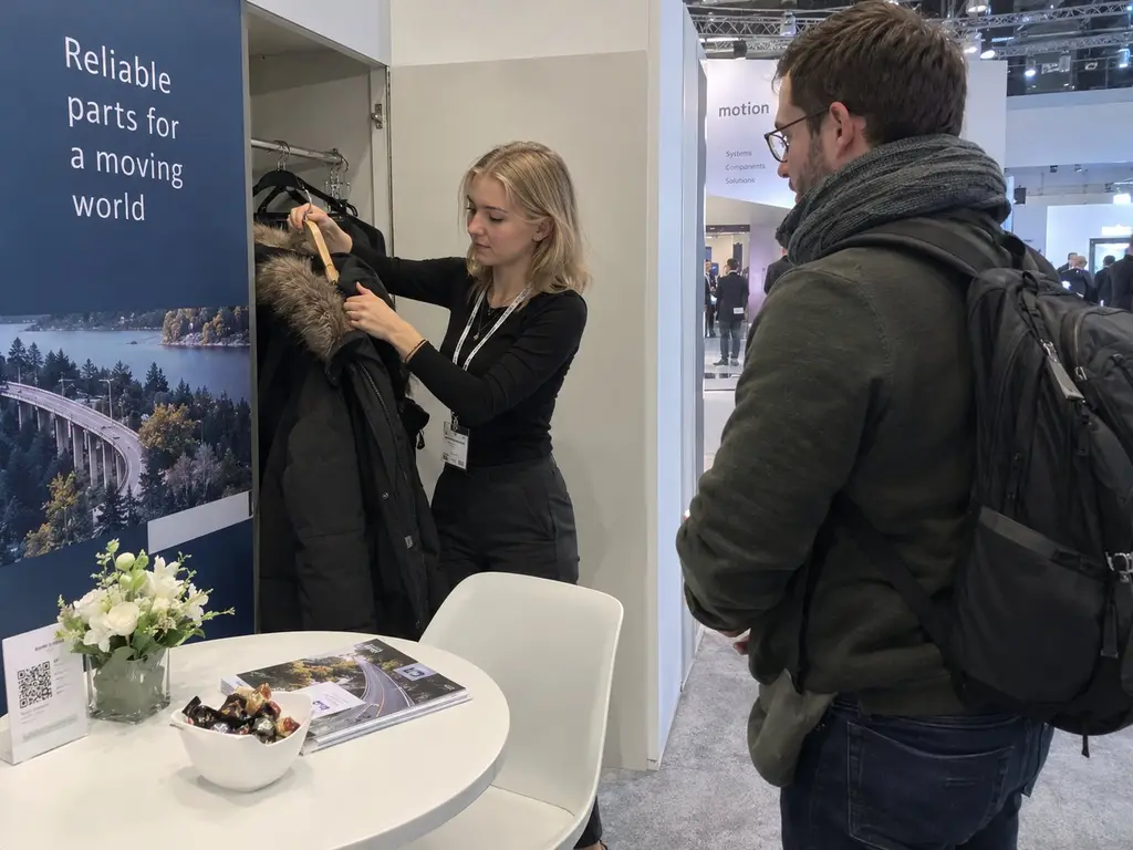 Standhostess hängt am Messestand die Winterjacke eines Besuchers in die Standgarderobe – Messepersonal in Berlin