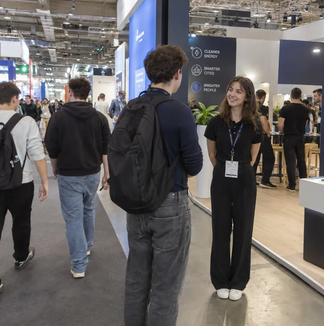 Hostess in a black polo shirt smiling as she greets a visitor at a trade fair stand