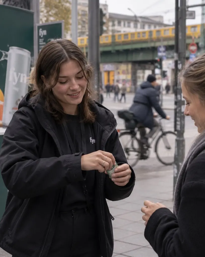 Promoterin gibt einer Passantin an einem Aktionsstand eine Probedose, dahinter die Hochbahn